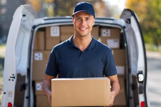 A smiling delivery man holds a cardboard box. He is standing in front of the open back of a delivery van that is filled with boxes.