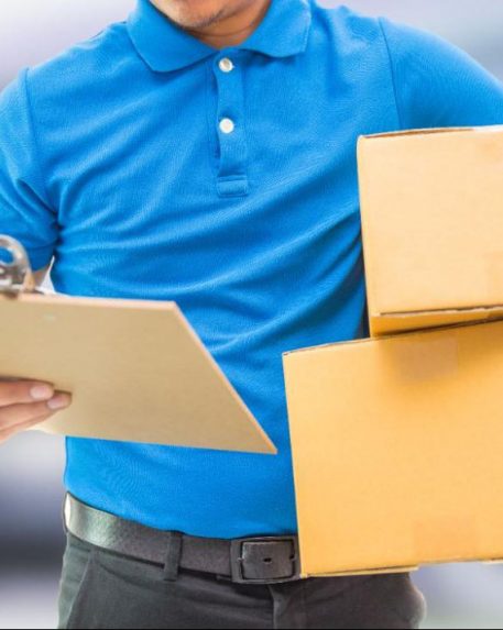 A man in a blue shirt looks at a clipboard in his right hand while carrying two cardboard boxes under his left arm.