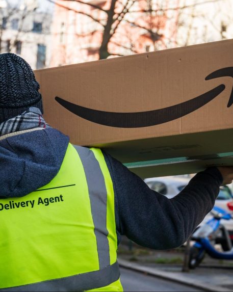 An Amazon delivery service provider in a reflective vest carries a large Amazon box in the city.