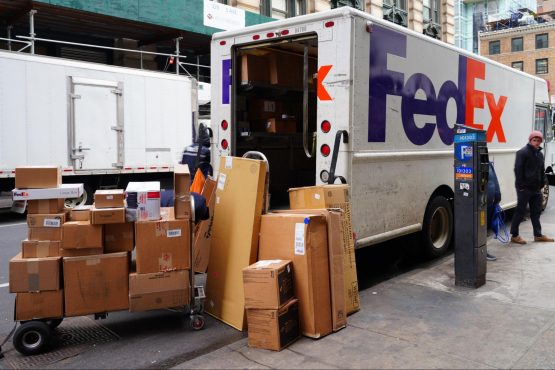 A FedEx truck is parked on a New York City street. Dozens of boxes have been unloaded and are sitting on the sidewalk or on a dolly.